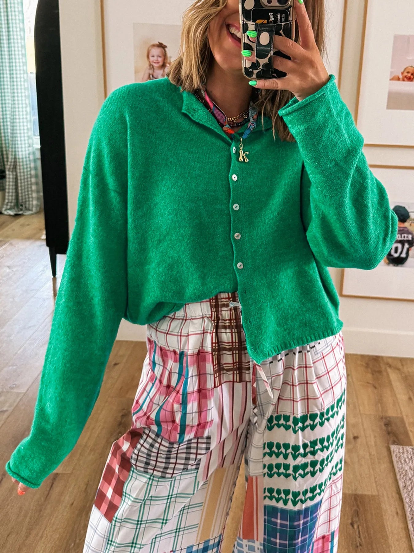 Person wearing a green cardigan over a colorful plaid dress, taking a mirror selfie.