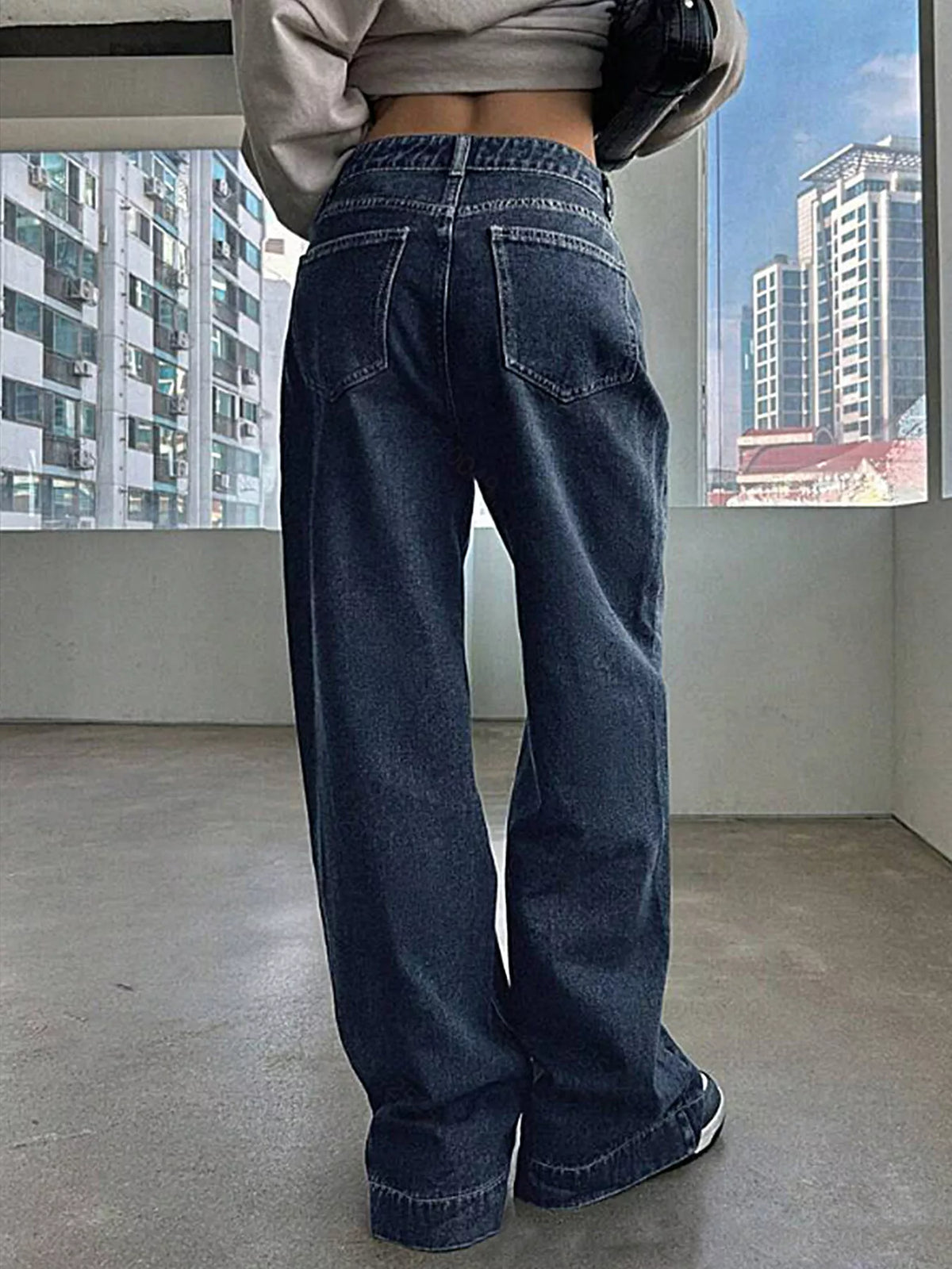 Person wearing blue jeans standing in a room with large windows showing cityscape.