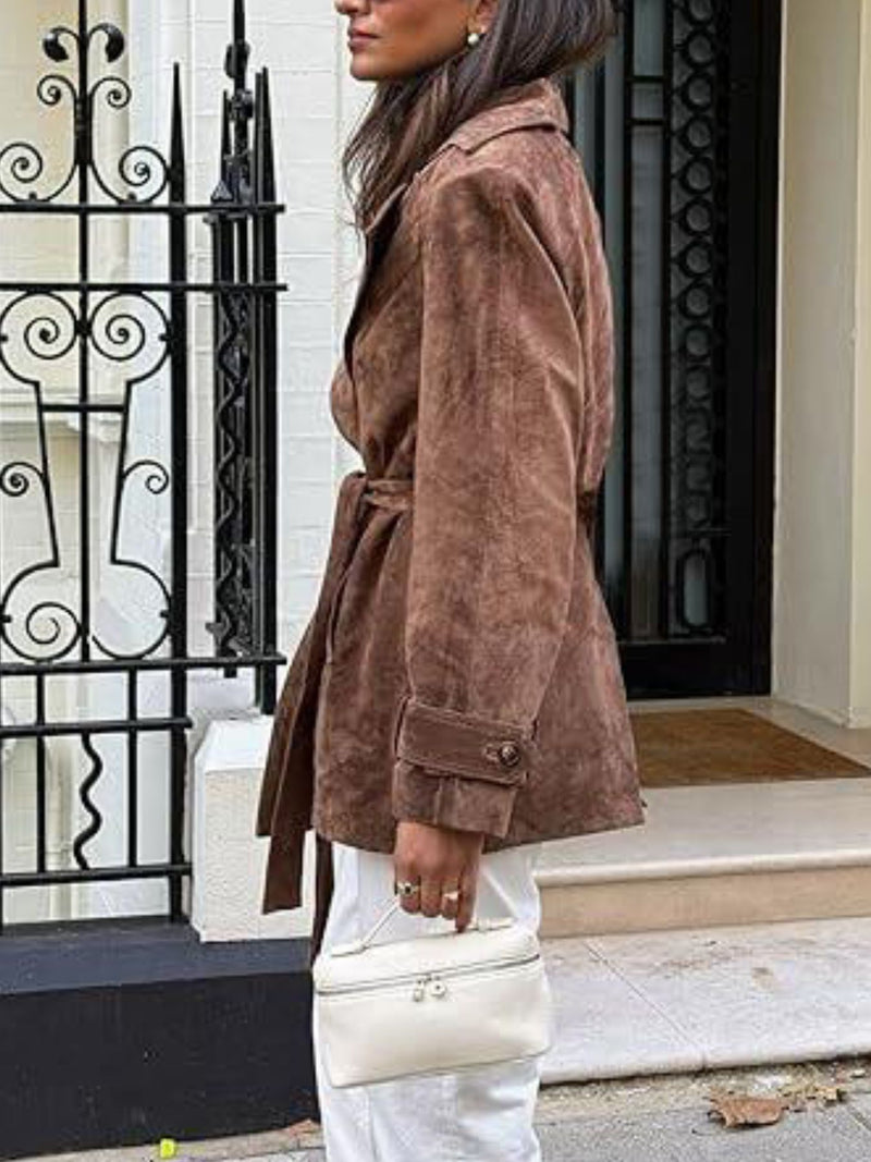 Person wearing a brown suede coat with a white bag, standing in front of a building entrance.