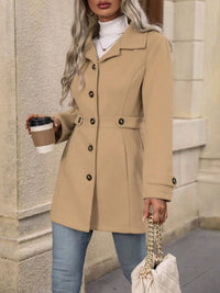 Woman wearing a beige coat holding a coffee cup and a handbag, standing against a stone wall.