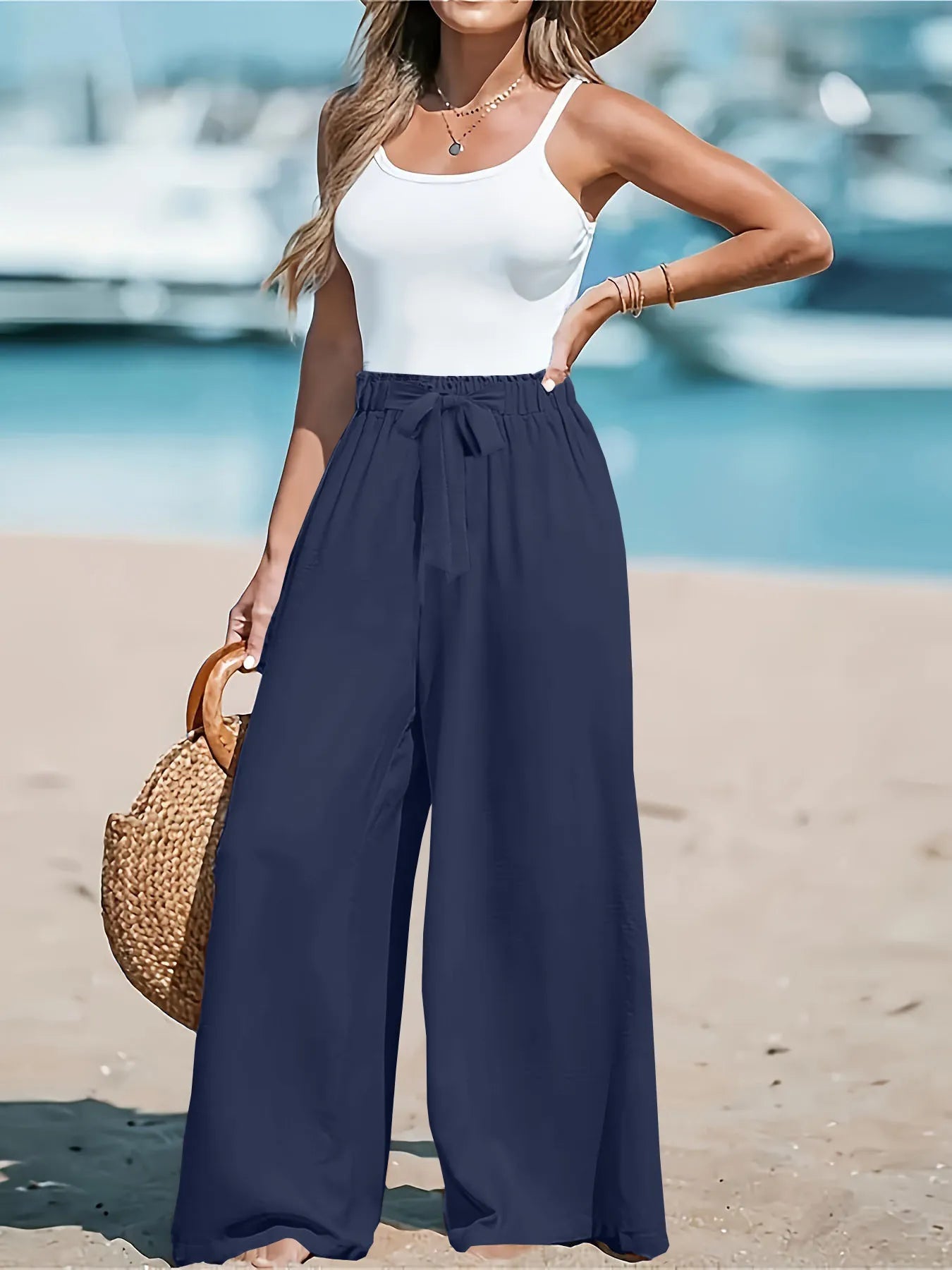 Woman in white top and navy wide-leg pants standing on a beach with boats in the background
