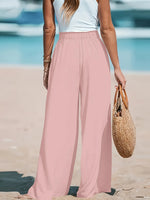 Person wearing pink wide-leg pants and a white top on a beach, holding a straw bag.