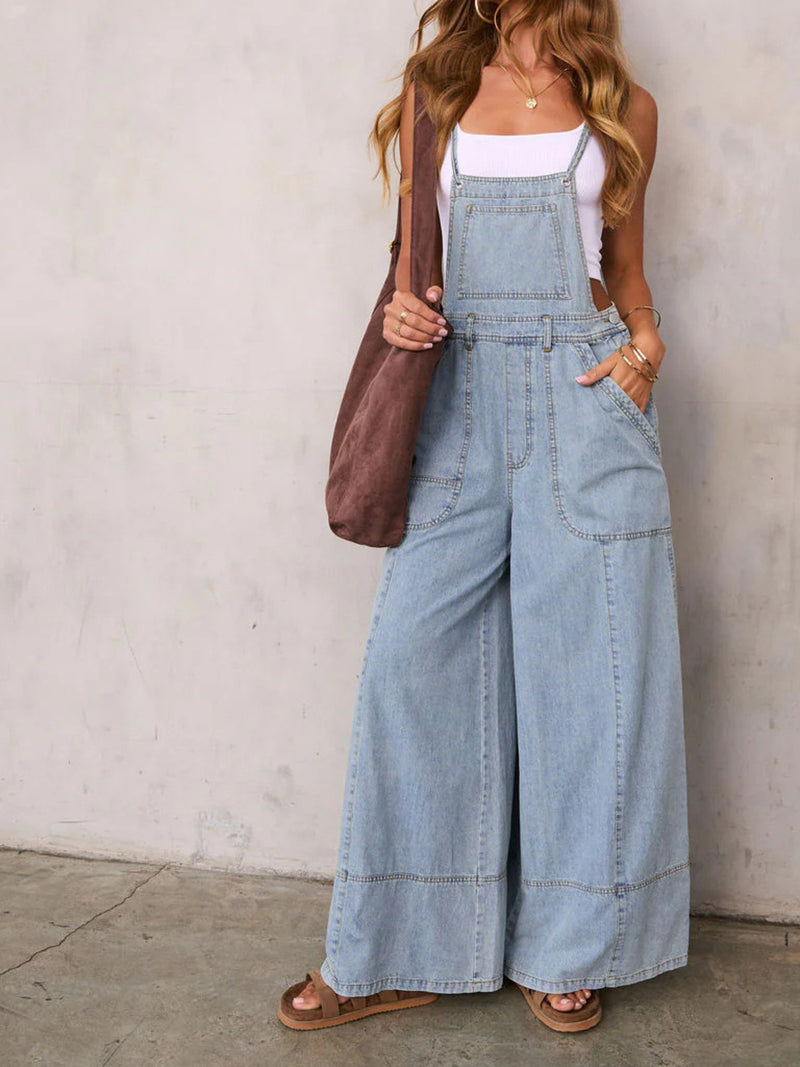 Woman wearing denim overalls with a white top and brown bag against a plain background