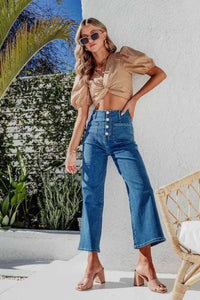 Woman in a beige top and blue jeans standing outdoors with plants and a chair in the background.