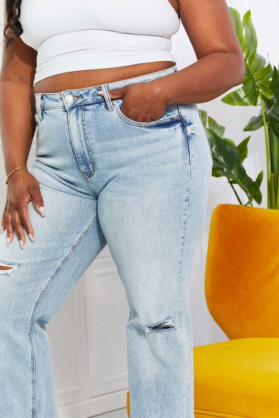Person wearing light blue jeans with a white top in a room with a yellow chair and green plant.