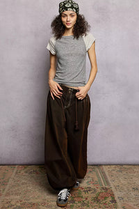 Woman wearing a gray t-shirt and brown wide-leg pants against a textured wall.