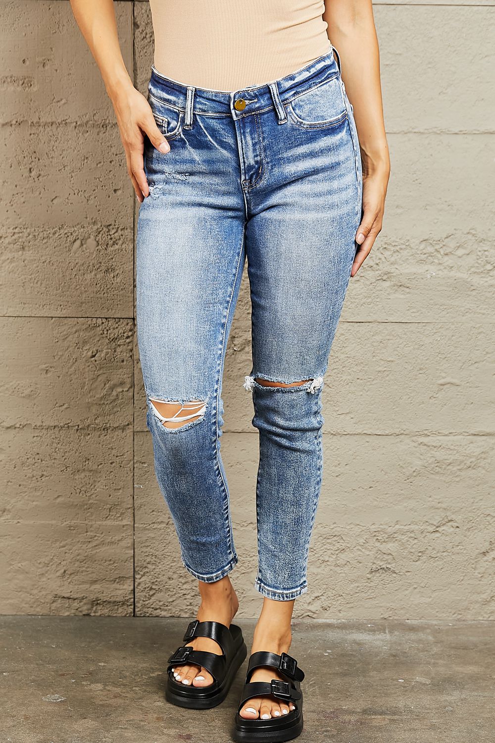 Person wearing distressed blue jeans and black sandals against a stone wall.