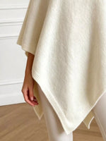 A close-up of a person wearing a soft cream-colored knit poncho draped over their shoulders, paired with light-colored leggings, in an indoor setting with a white paneled wall and light wood floor.