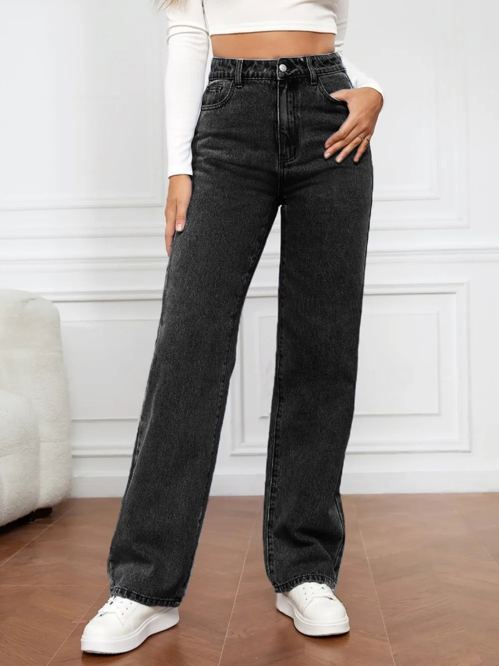 Person wearing black high-waisted jeans and white sneakers in a minimalistic room.