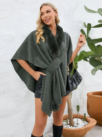 Woman wearing a dark green poncho with fur trim against a white wall with plants.