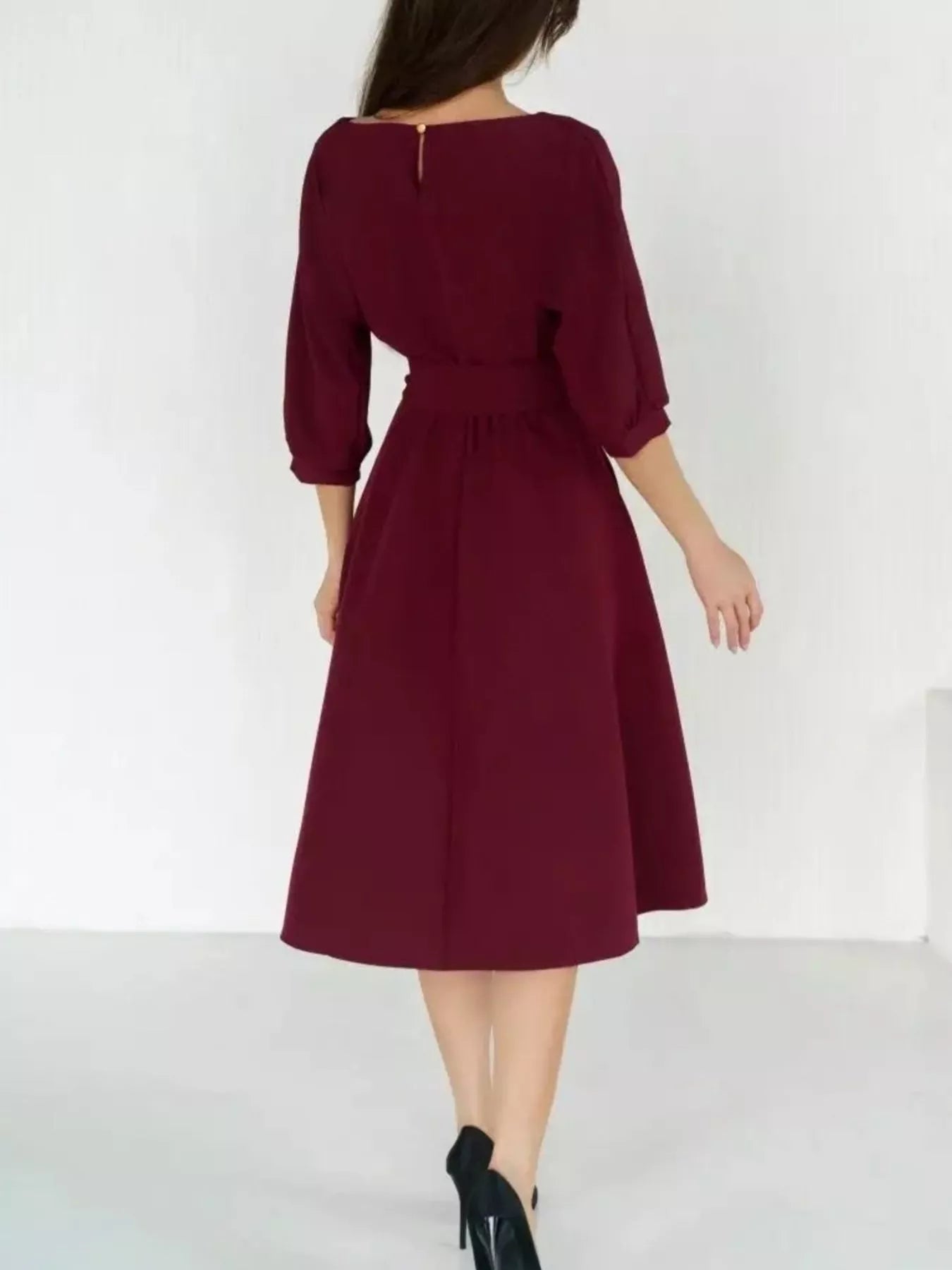 A woman wearing a knee-length burgundy dress with three-quarter sleeves and a gathered waist, shown from the back against a plain white studio background.