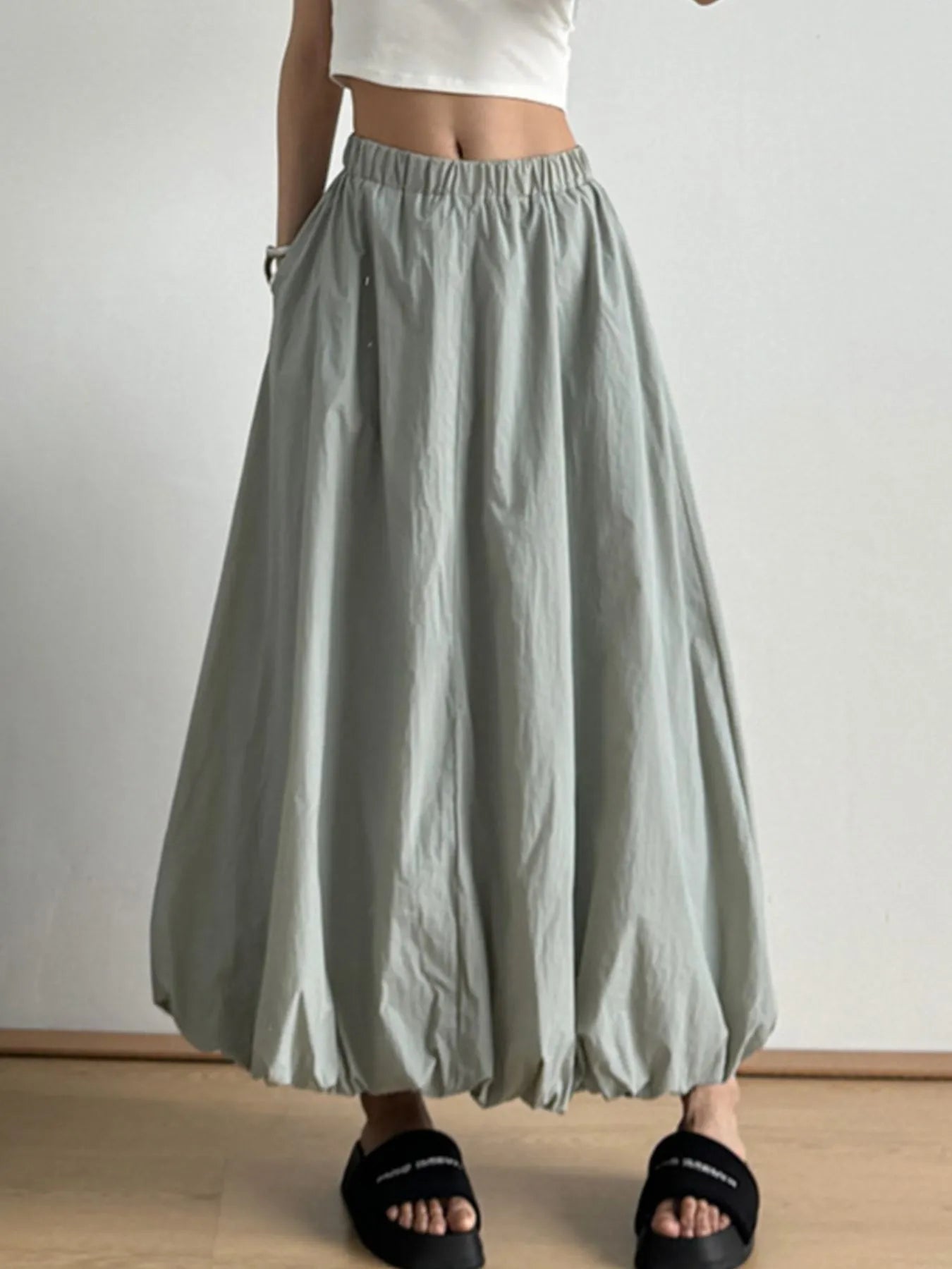 Person wearing a light gray skirt with a white top and black sandals on a plain background