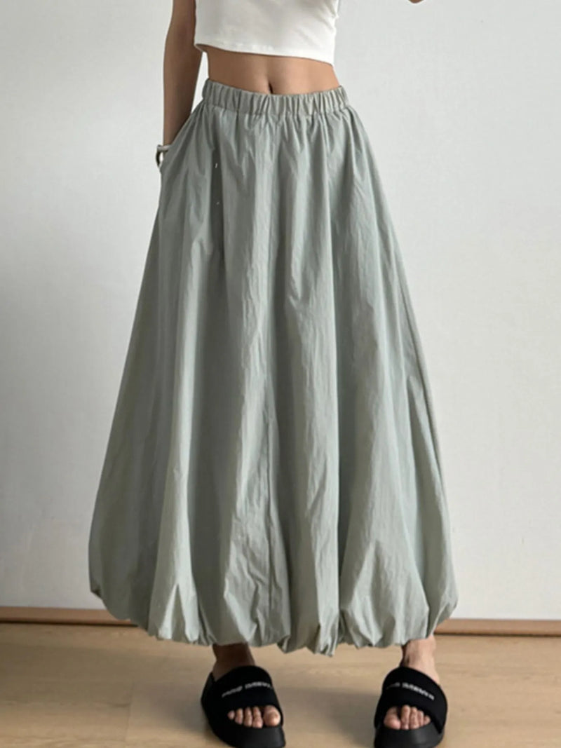 Person wearing a light gray skirt with a white top and black sandals on a plain background