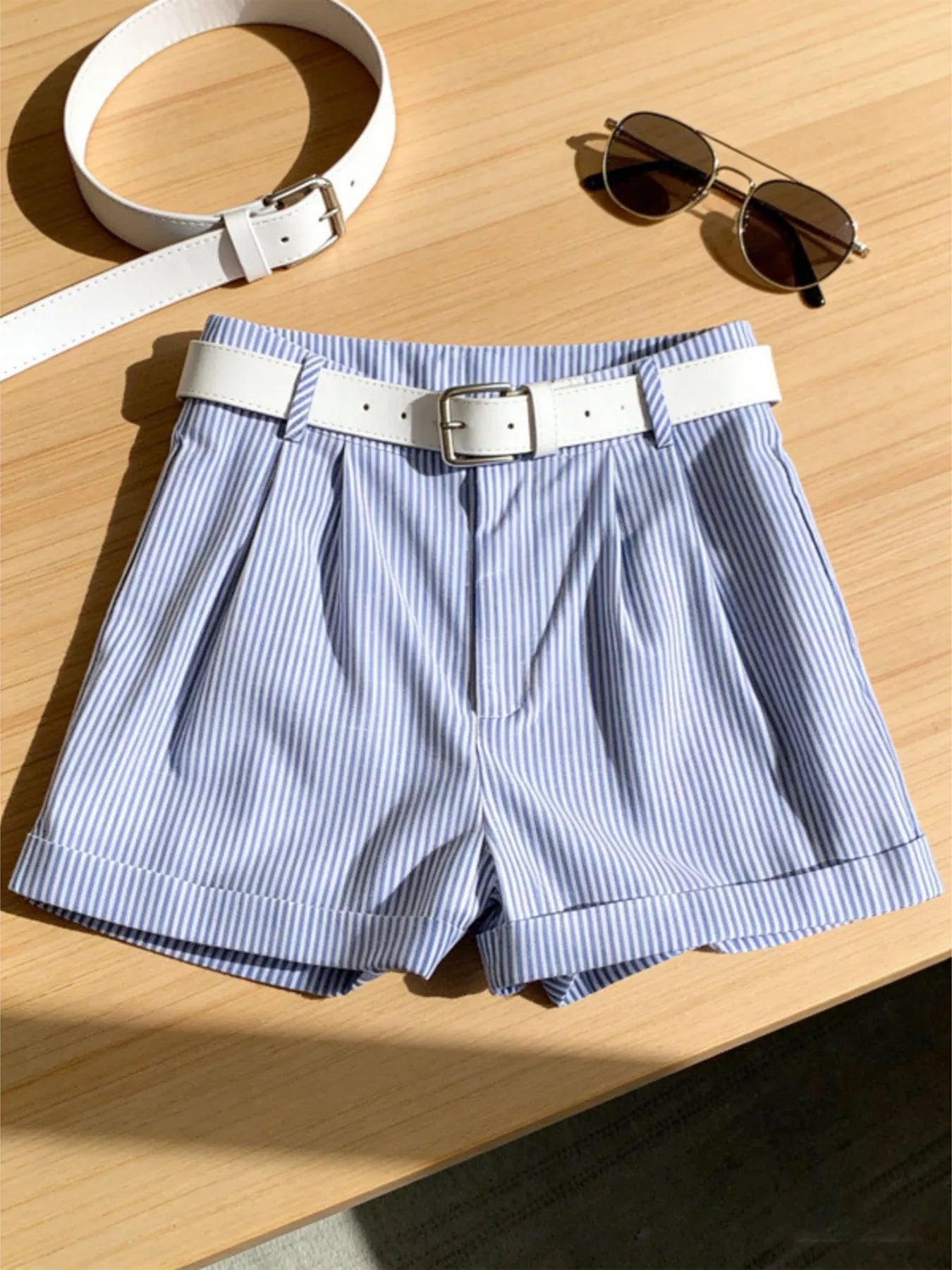 Blue striped shorts with a white belt on a wooden surface, accompanied by a white belt and sunglasses.