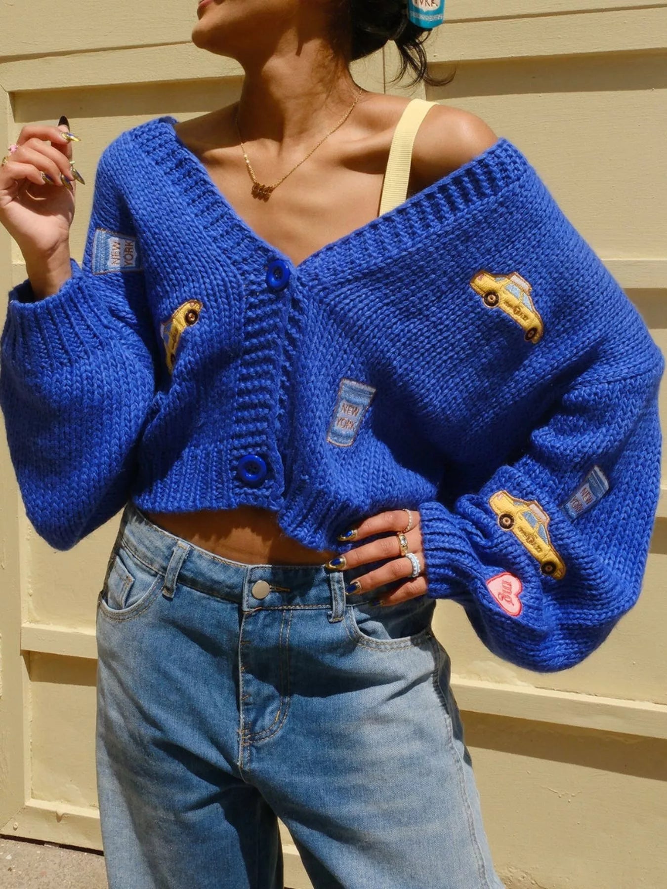 Person wearing a blue knitted cardigan with patches in front of a beige wall.