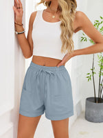 Woman wearing a white crop top and light blue shorts against a white wall with a plant.