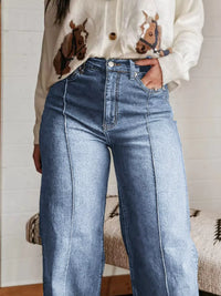 Person wearing a cream sweater with horse designs and blue jeans indoors.