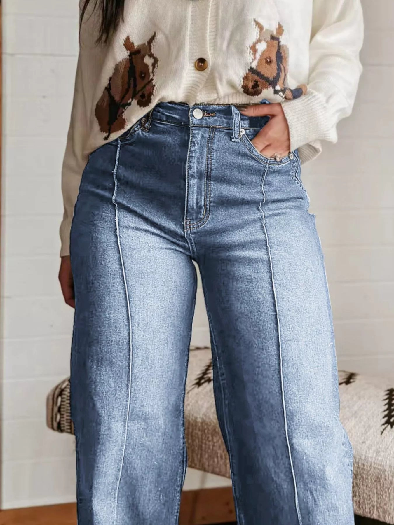 Person wearing a cream sweater with horse designs and blue jeans indoors.