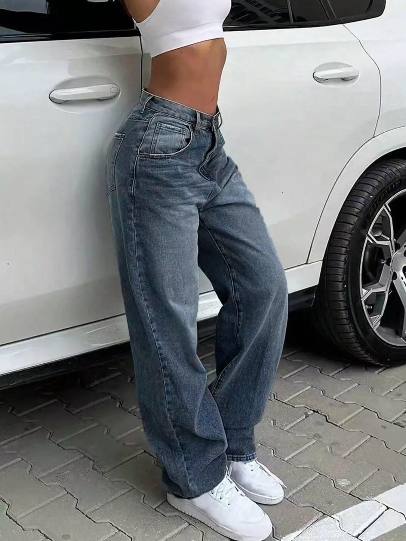 Person wearing blue jeans and white sneakers leaning against a white car.