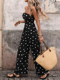 Woman wearing a black polka dot jumpsuit holding a straw bag against a textured wall.