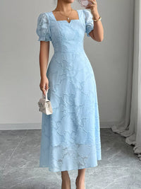 Woman wearing a light blue lace dress holding a small white handbag indoors.