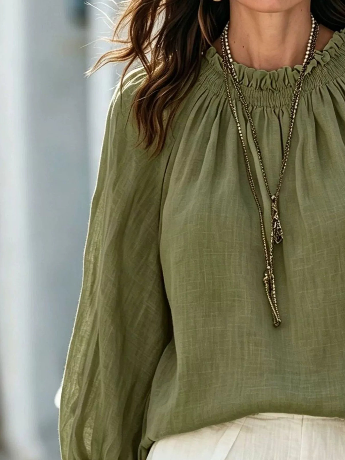 A close-up of a woman wearing a loose, long-sleeve olive green blouse with a gathered smocked neckline, layered with long silver chain necklaces, and paired with white bottoms.