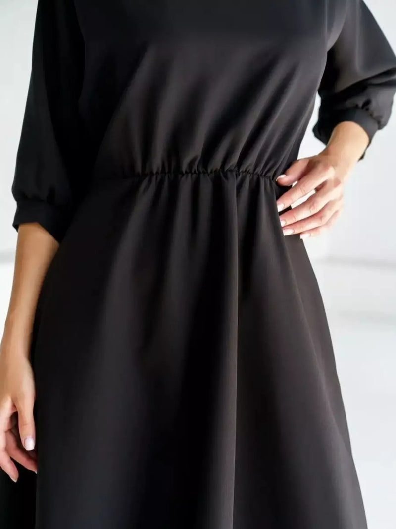 A close-up of a black dress worn by a model, showing the three-quarter sleeves, gathered waist, and flowing skirt against a light background.