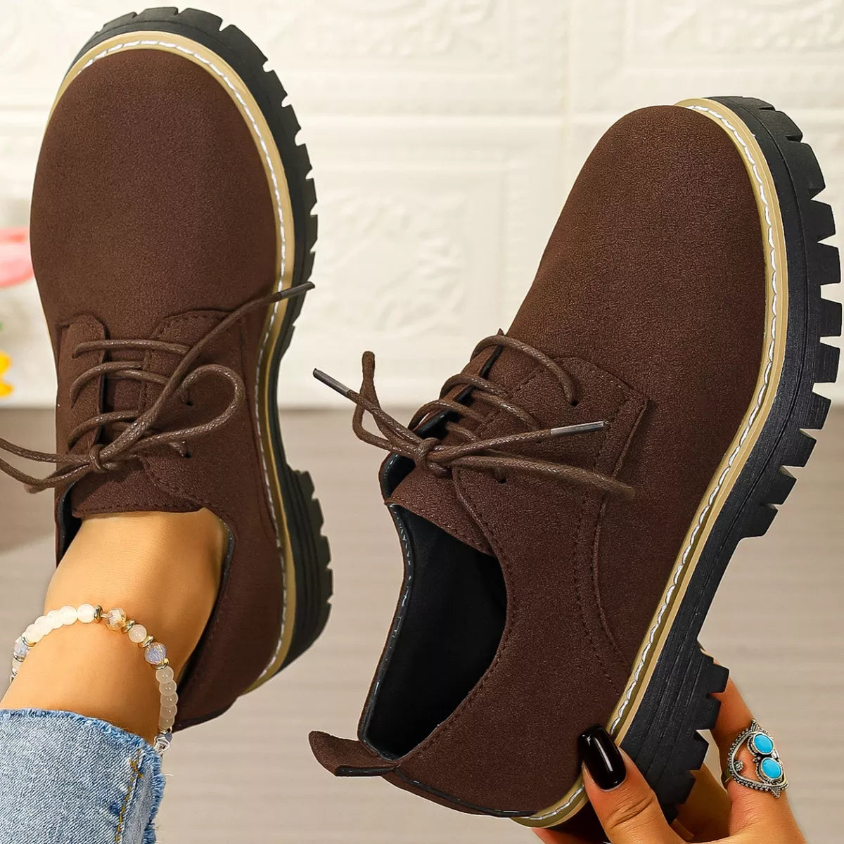 Brown suede shoes with black soles held by a person against a neutral background