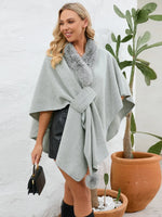 Woman wearing a light gray poncho with a fur collar, standing next to a potted plant indoors.