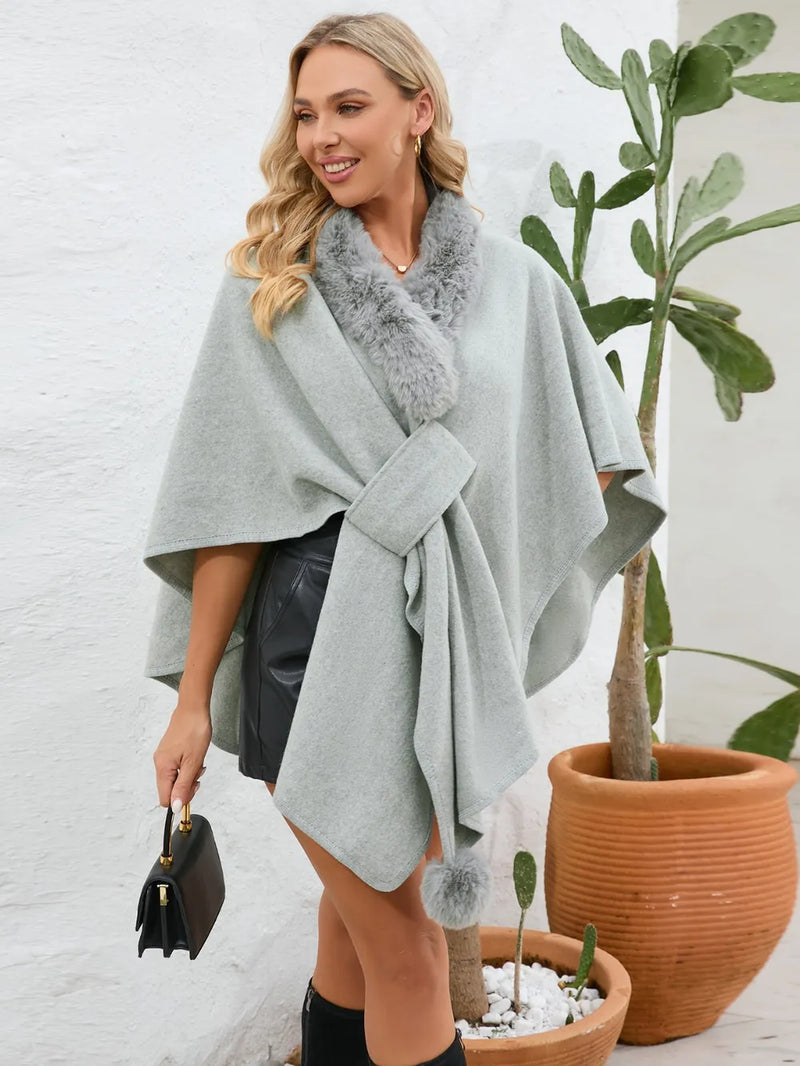 Woman wearing a light gray poncho with a fur collar, standing next to a potted plant indoors.