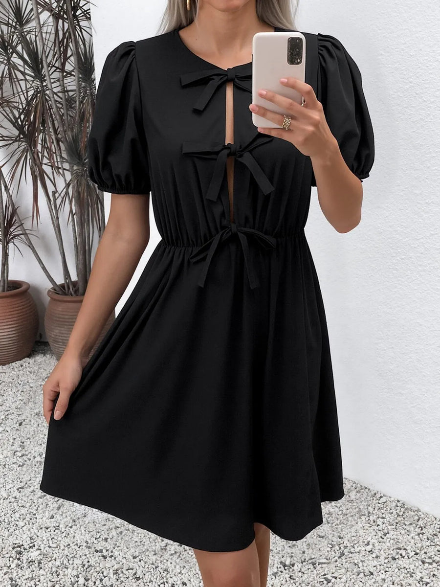 Woman wearing a black dress with puff sleeves, standing against a white wall.