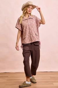 Woman wearing a patterned shirt and brown pants with a beige hat against a pink background