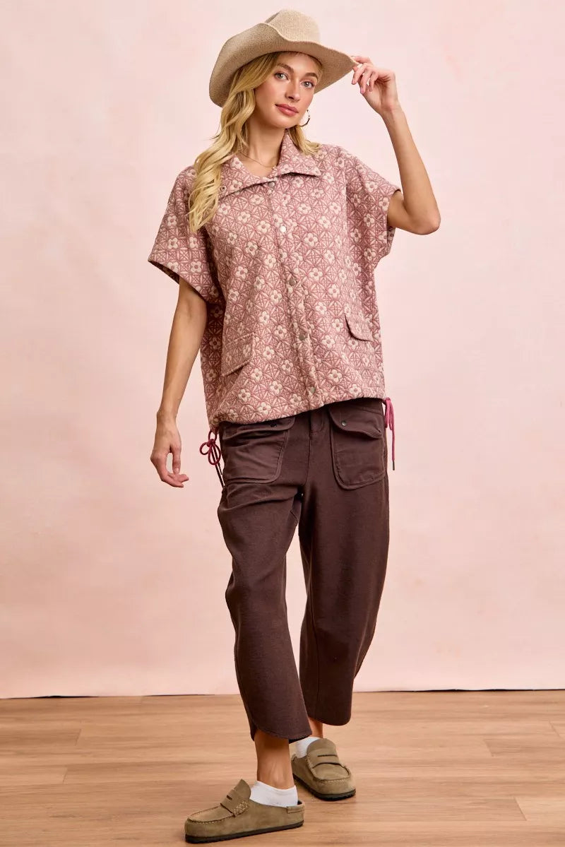 Woman wearing a patterned shirt and brown pants with a beige hat against a pink background