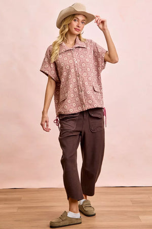 Woman wearing a patterned shirt and brown pants with a beige hat against a pink background