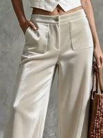 A close-up of a model wearing high-waisted, wide-leg trousers in a shimmering champagne beige fabric. The pants feature a button closure, a single front patch pocket, and a relaxed, flowing silhouette.