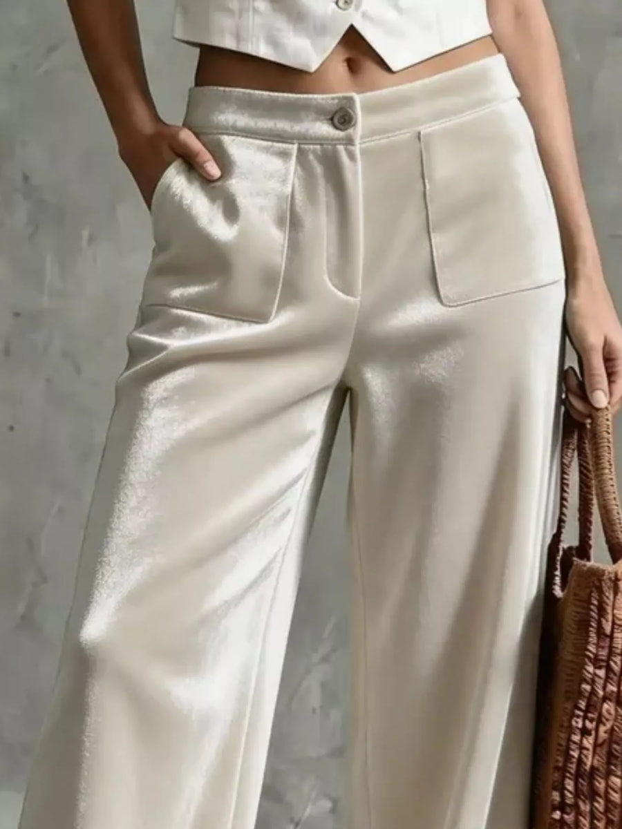 A close-up of a model wearing high-waisted, wide-leg trousers in a shimmering champagne beige fabric. The pants feature a button closure, a single front patch pocket, and a relaxed, flowing silhouette.