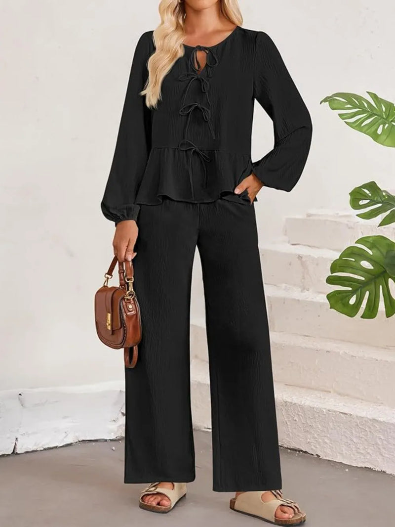 Woman wearing a black outfit with wide-leg pants and a brown handbag, standing against a white wall with green leaf patterns.