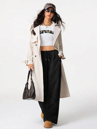 Woman wearing a long beige coat, white crop top, black pants, and brown boots on a white background
