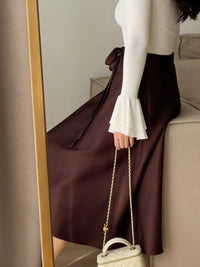 Person wearing a white top and brown skirt holding a cream-colored handbag in front of a mirror.