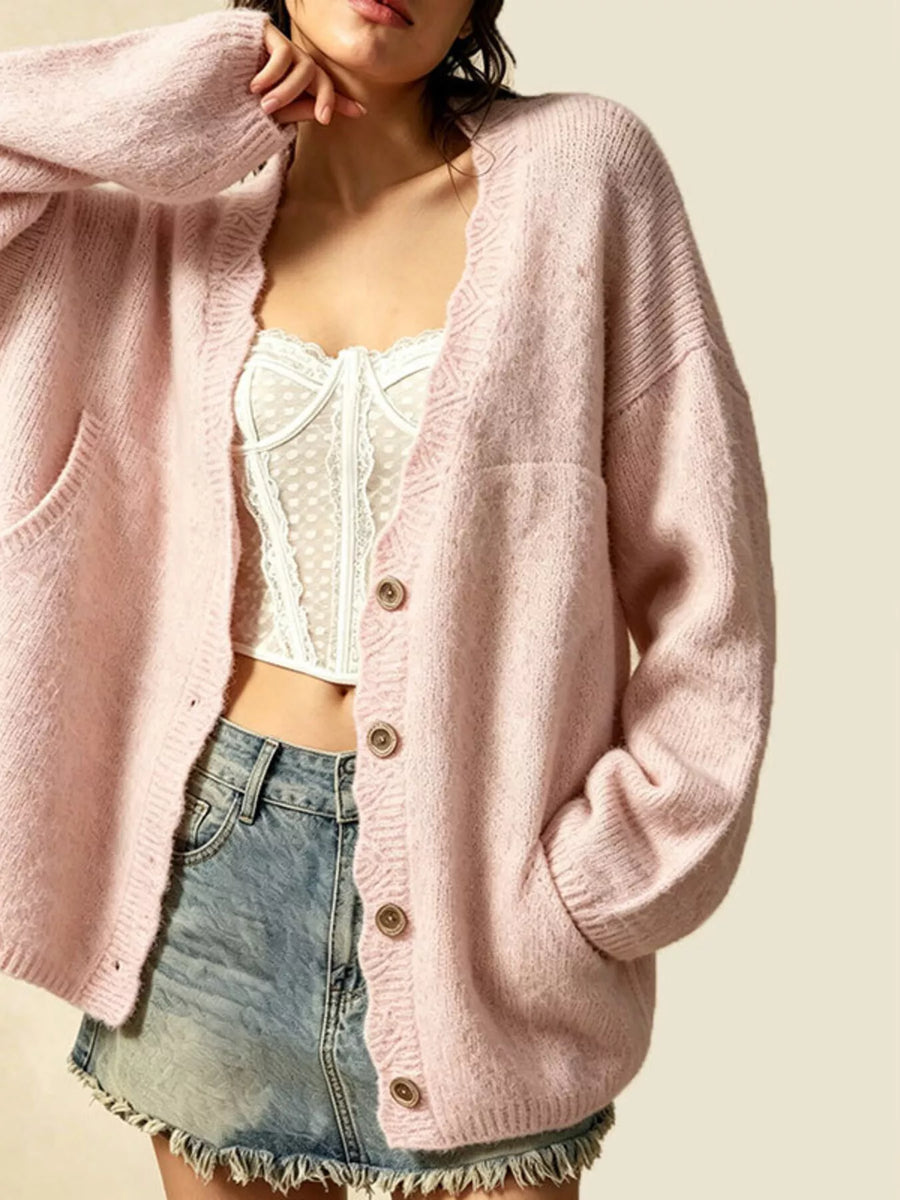 A woman wearing a soft pink open-front knit cardigan with buttons, layered over a white lace-trim camisole and paired with a frayed denim mini skirt, against a plain light beige background.