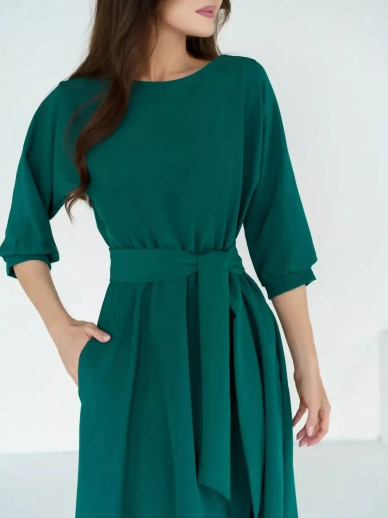 A woman wearing a dark green midi dress with three-quarter sleeves, a round neckline, and a matching fabric tie belt at the waist, shown against a plain white background.