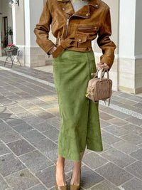 Person wearing a brown jacket and green skirt holding a small handbag on a stone pavement.