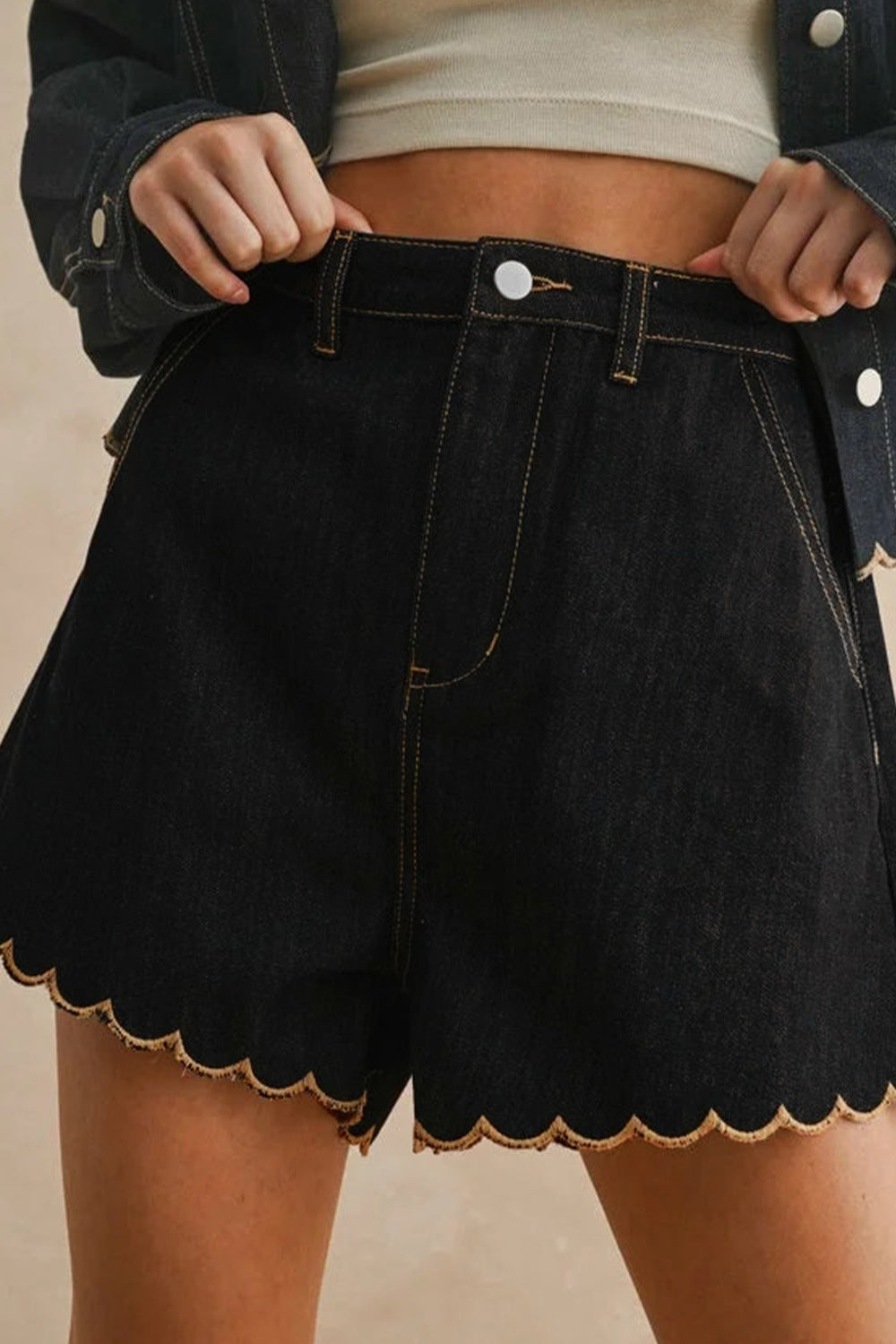 Person wearing black denim shorts with scalloped hem on a beige background