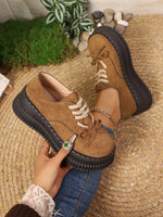 Brown suede shoes with black soles worn by a person on a textured surface with plants in the background