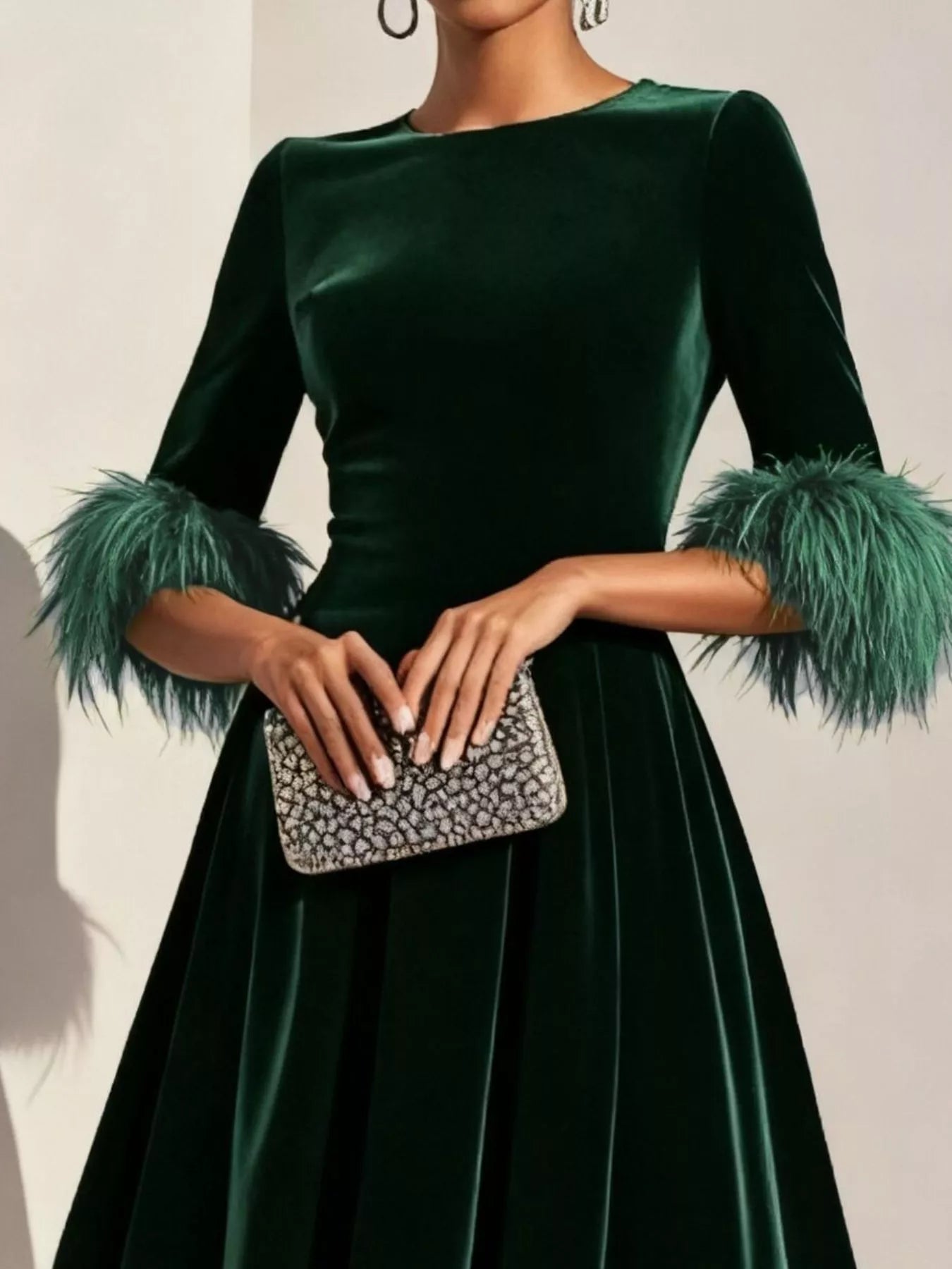 Green dress with feathered sleeves held by a hand with a clutch against a neutral background