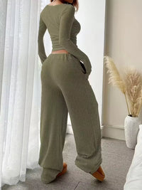 Person wearing a green ribbed outfit standing in a room with white curtains and a vase.