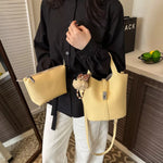 A person wearing a black blouse and white pants carries two light yellow pebbled leather bags in an indoor room. One is a small zip pouch with a black tie, and the other is a structured shoulder bag with a long strap and a small plush keychain charm.