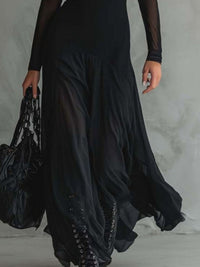 Person wearing a long black dress with sheer sleeves and holding a matching bag against a neutral background