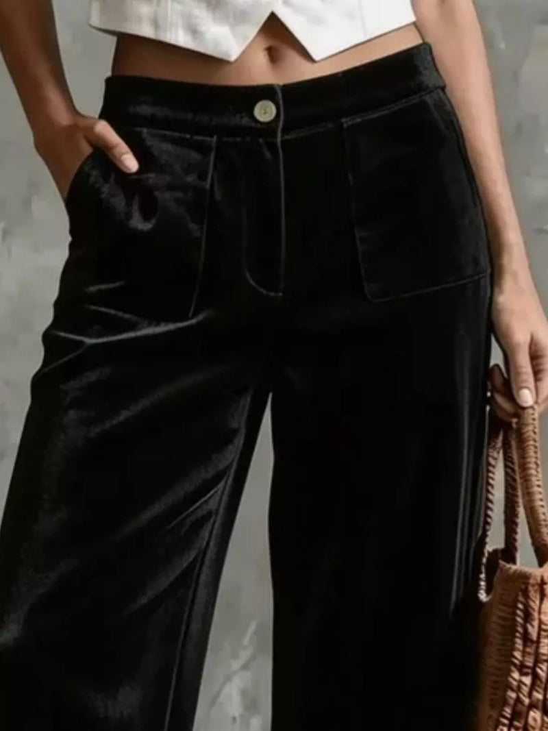 A close-up of a person wearing black wide-leg velvet pants with a high waist, front button closure, and flap pockets, paired with a white cropped top and holding a woven brown bag.