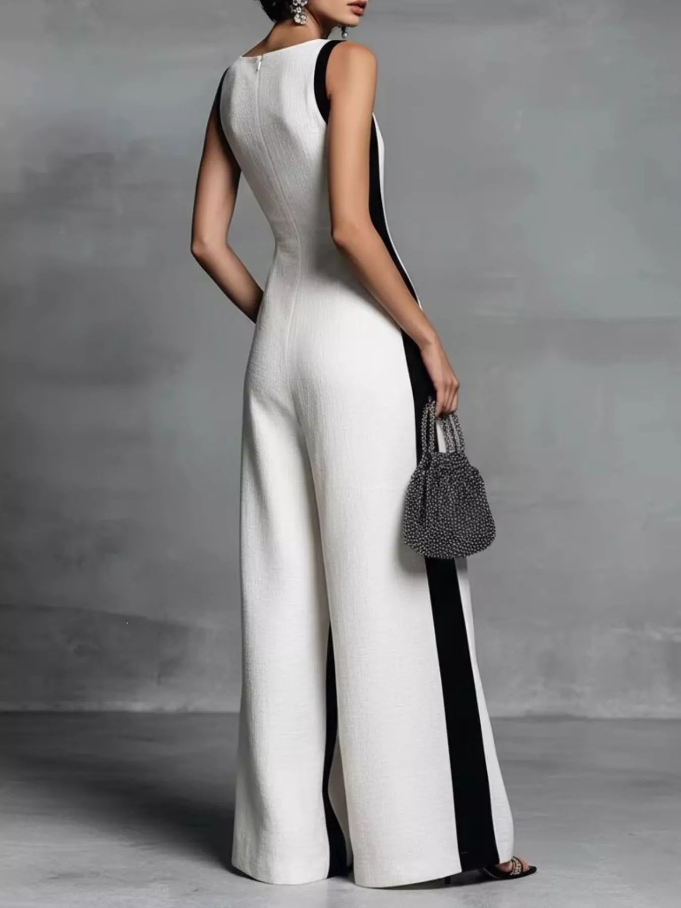 Woman wearing a white and black jumpsuit holding a handbag against a gray background